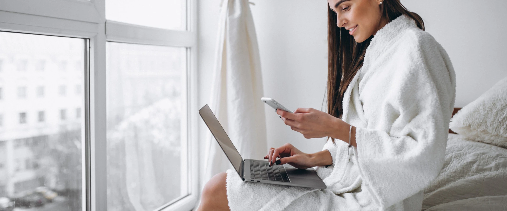Femme en peignoir utilisant un téléphone et un ordinateur portable près d'une fenêtre.