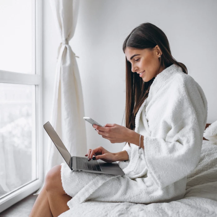 Femme en peignoir utilise un téléphone et un ordinateur portable sur un lit.