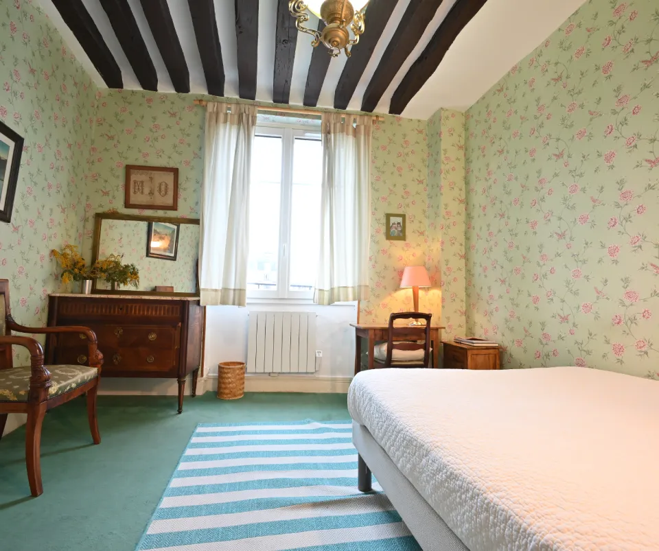 Habitación vintage con papel tapiz floral, cama blanca y muebles de madera. Ventana iluminada.