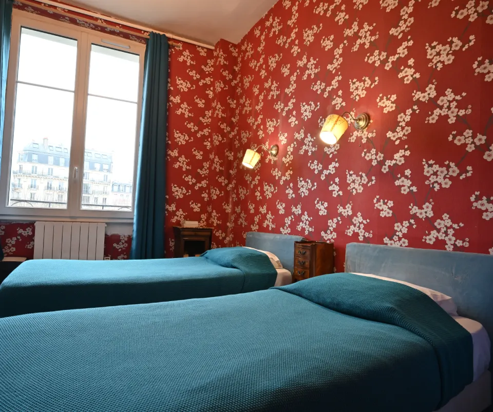 Habitación con dos camas, papel tapiz floral rojo, ventanas grandes y cubrecamas azules.