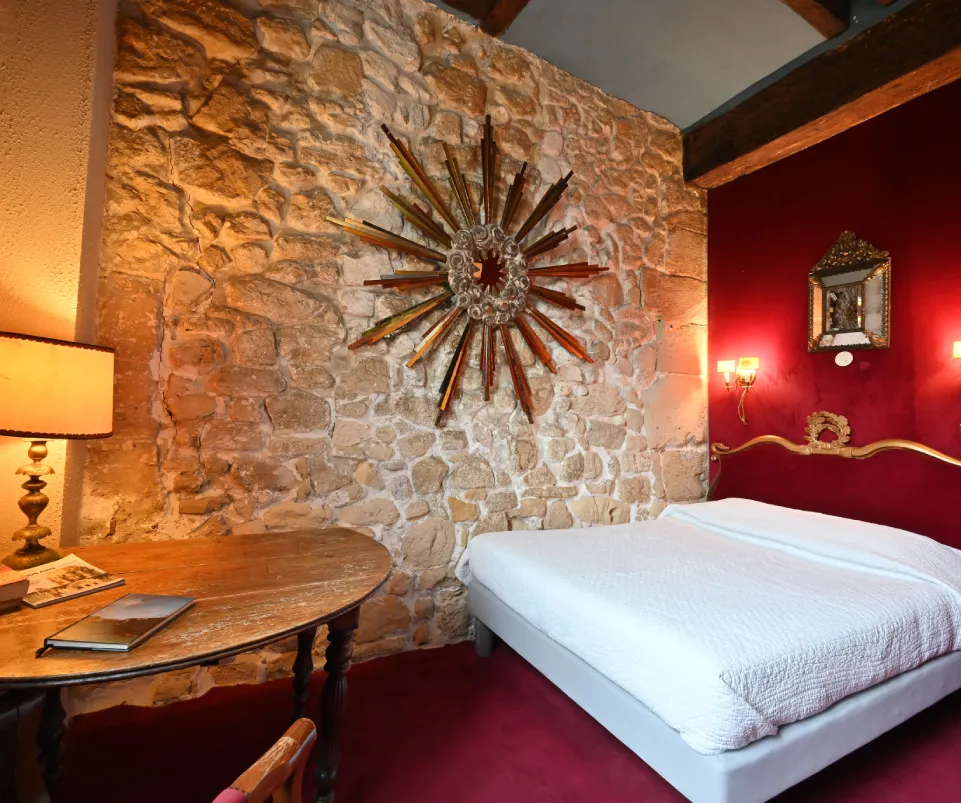 Habitación con paredes de piedra y roja, cama blanca, lámparas y mesa redonda de madera.