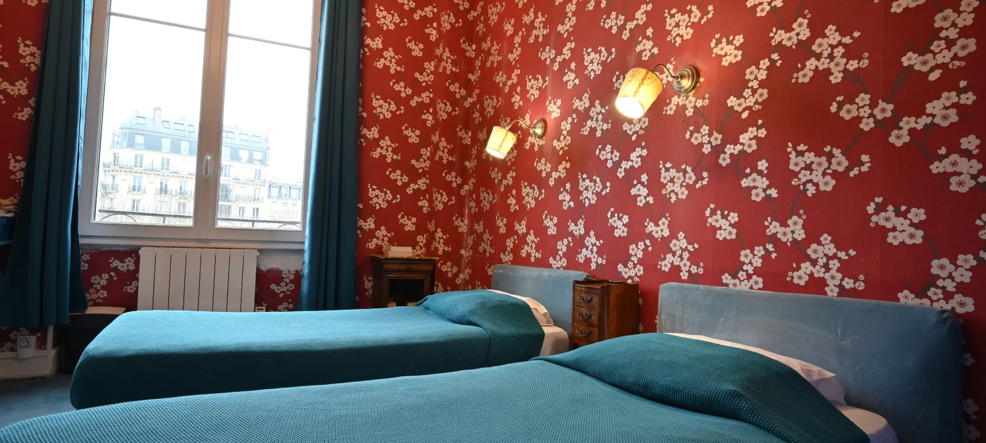 Habitación con dos camas, pared roja floral y ventana con vista urbana.