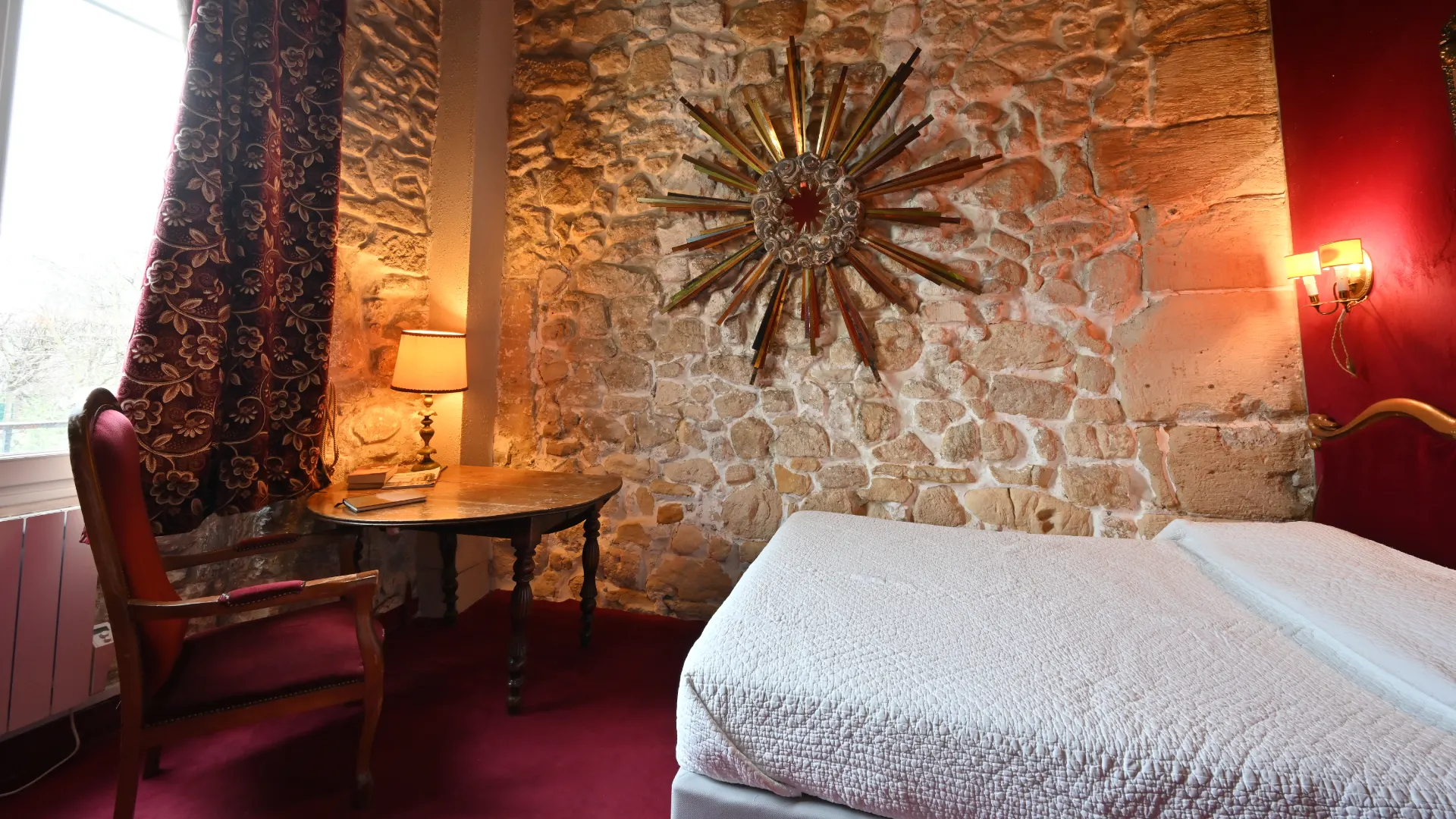 Habitación con piedra vista, cama blanca, mesa, lámpara y adorno en forma de sol en la pared.