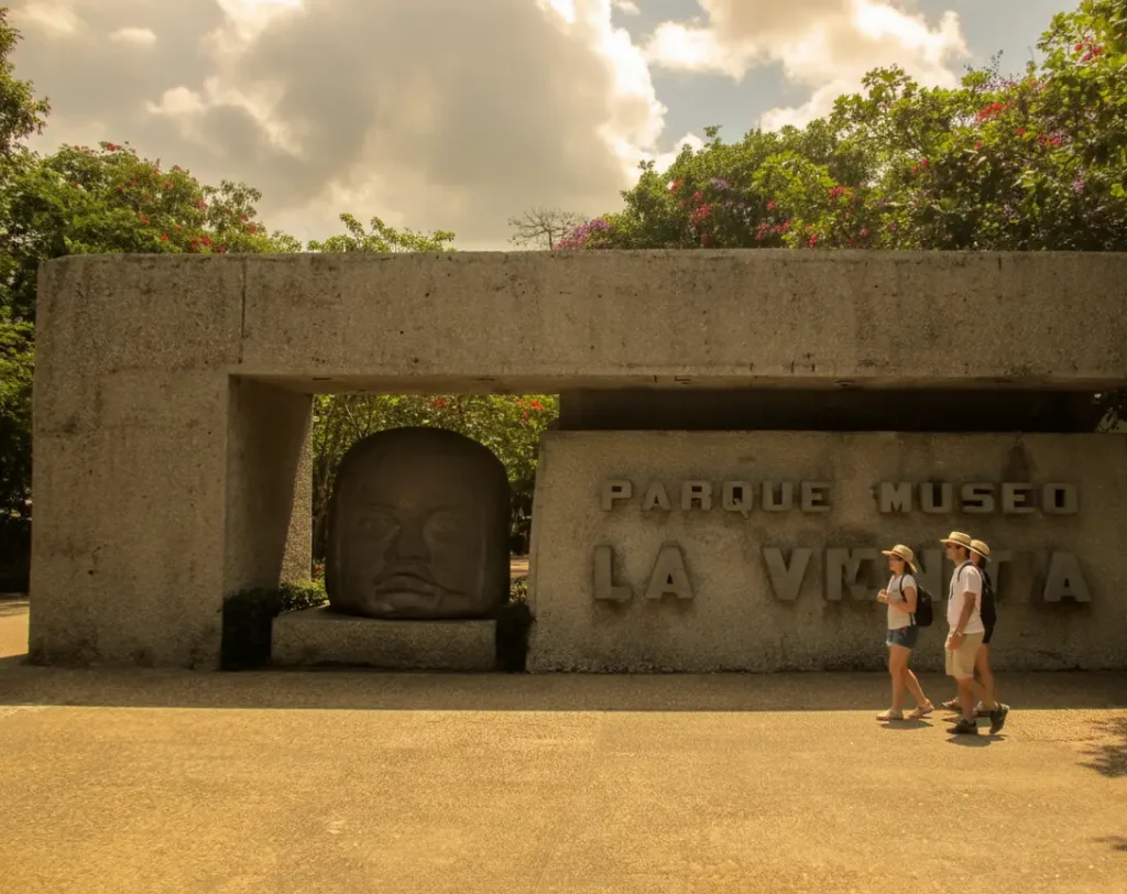 Entrada al Parque Museo La Venta con cabeza olmeca y tres personas caminando. Cielo nublado y vegetaci&oacute;n alrededor.
