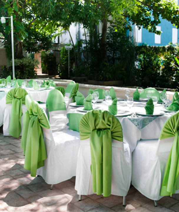 Mesas de banquete al aire libre decoradas con manteles blancos y cubiertas de silla verdes, rodeadas de vegetaci&oacute;n.