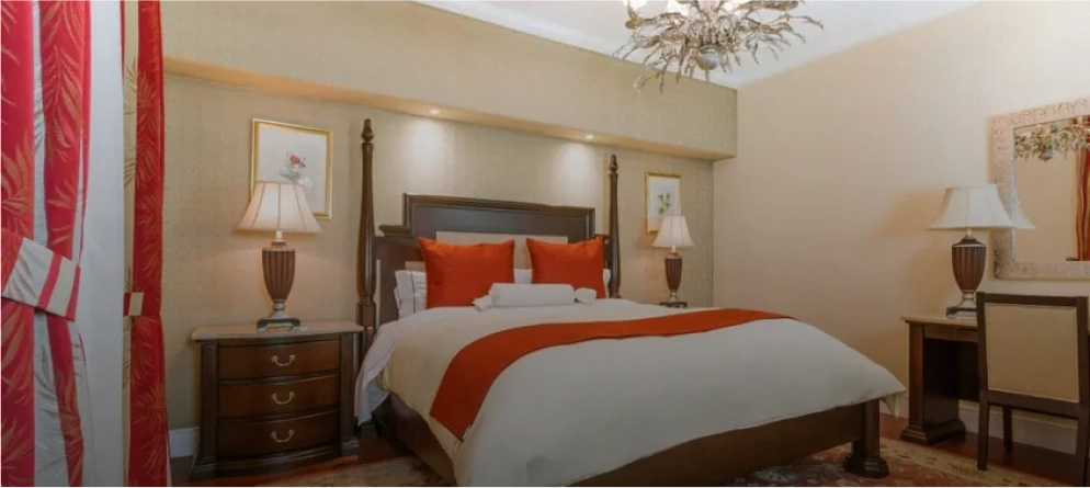 Habitaci&oacute;n con cama grande, cojines naranjas, l&aacute;mparas de mesa y un espejo en la pared. Decoraci&oacute;n elegante.