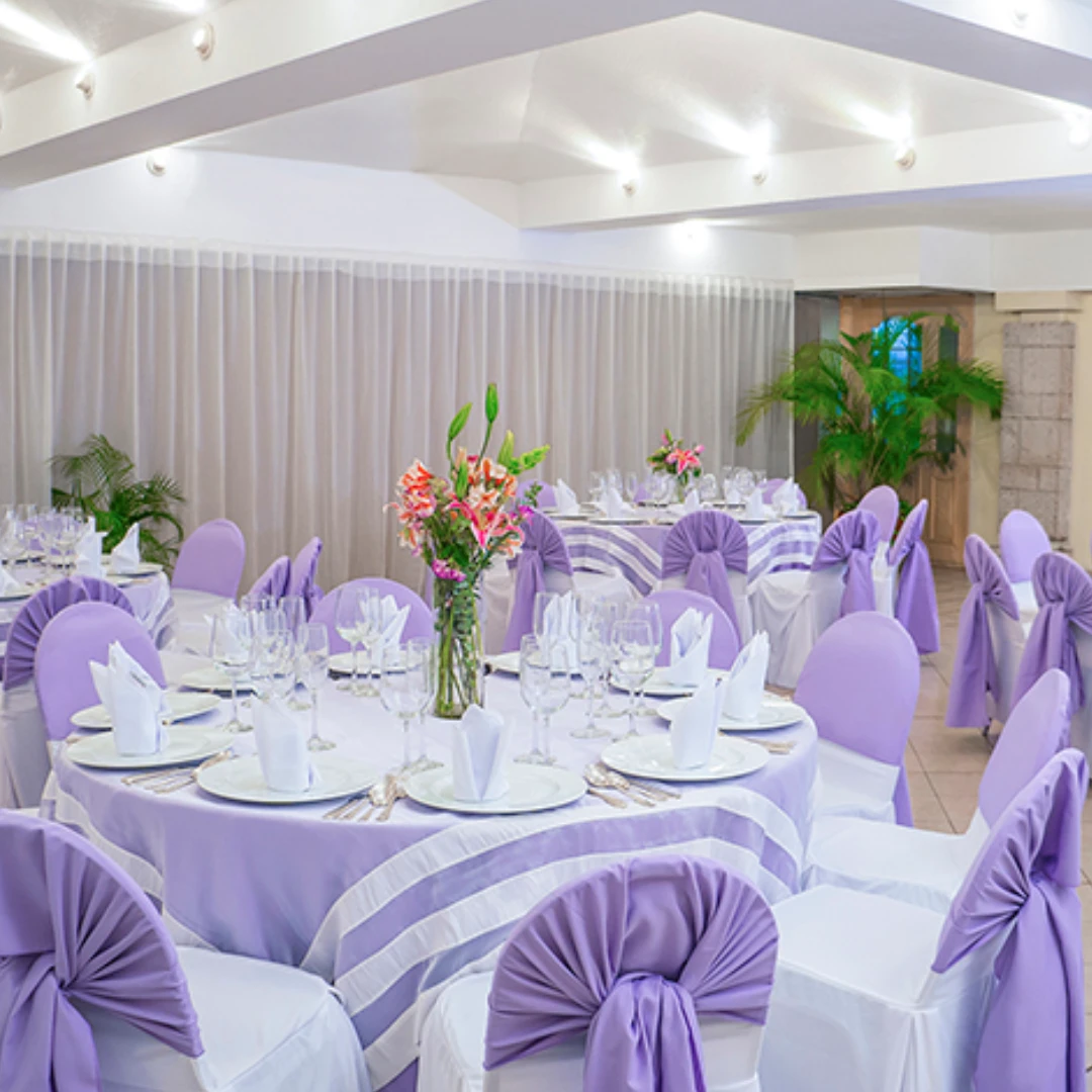 Sal&oacute;n decorado para evento con mesas redondas, manteles lilas, sillas forradas y centro de mesa floral.