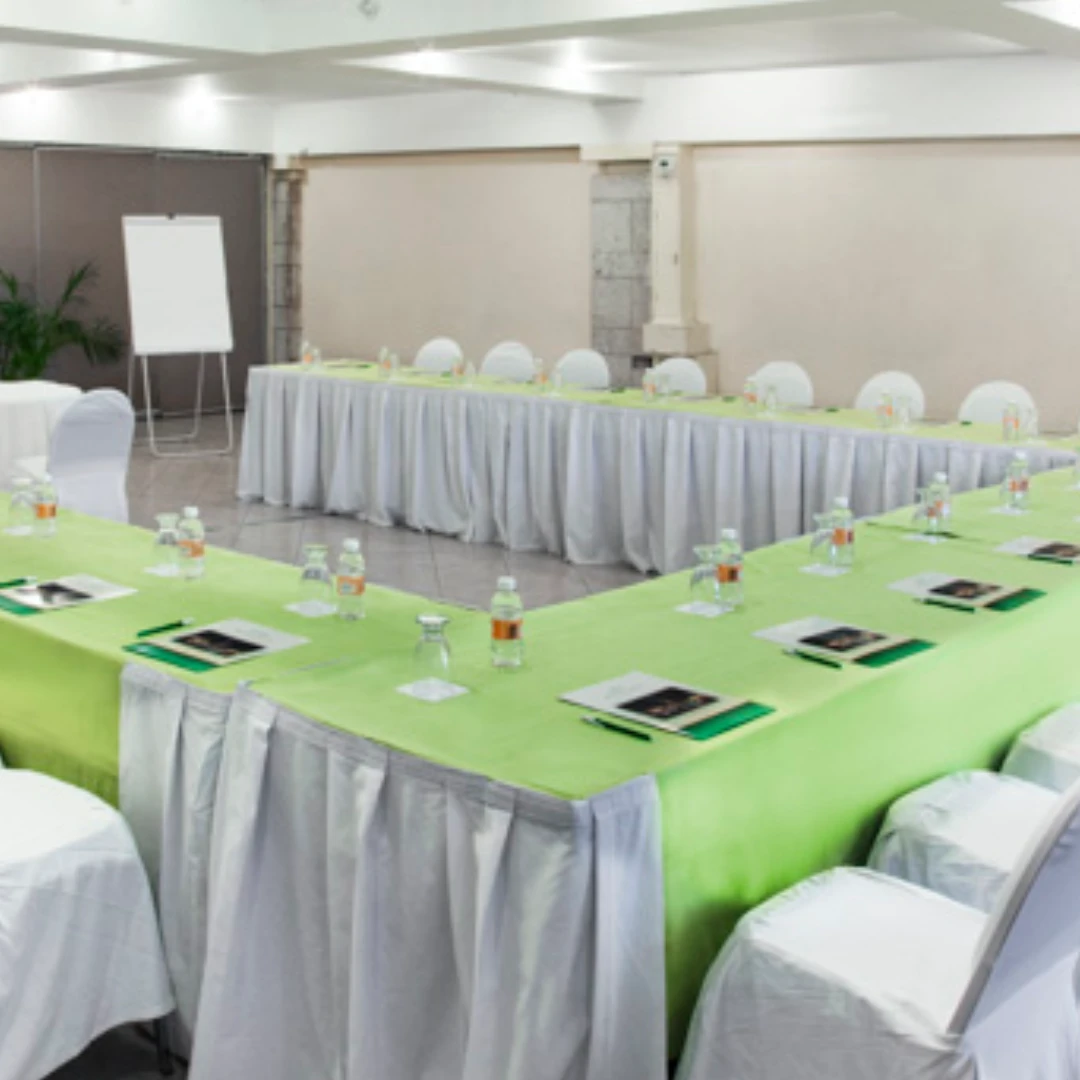 Sala de conferencias con mesas dispuestas en forma de U, sillas blancas, botellas de agua y material de escritura.