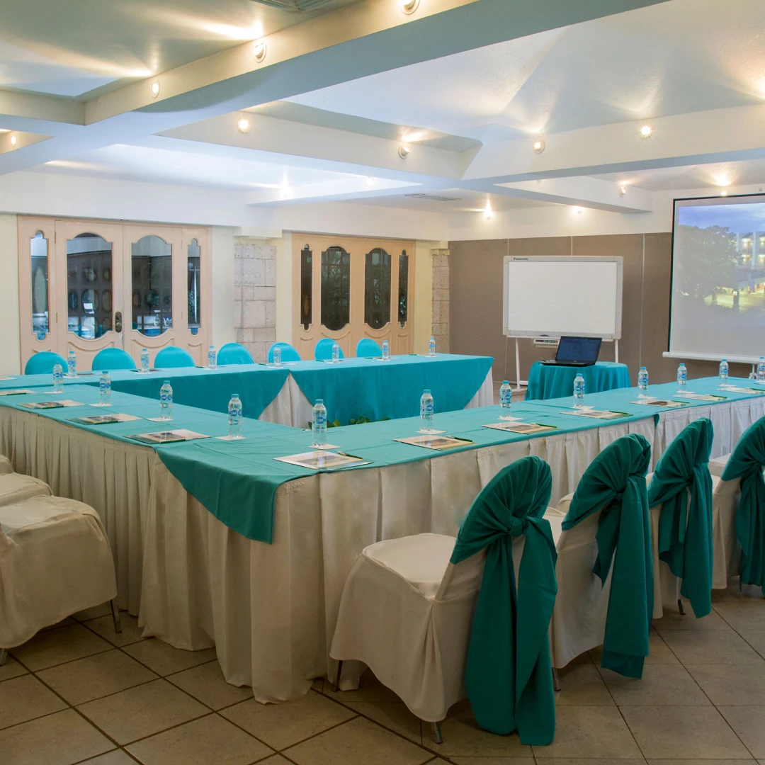 Sala de conferencias con mesa en U, sillas decoradas de verde, botellas de agua y proyector con pantalla.