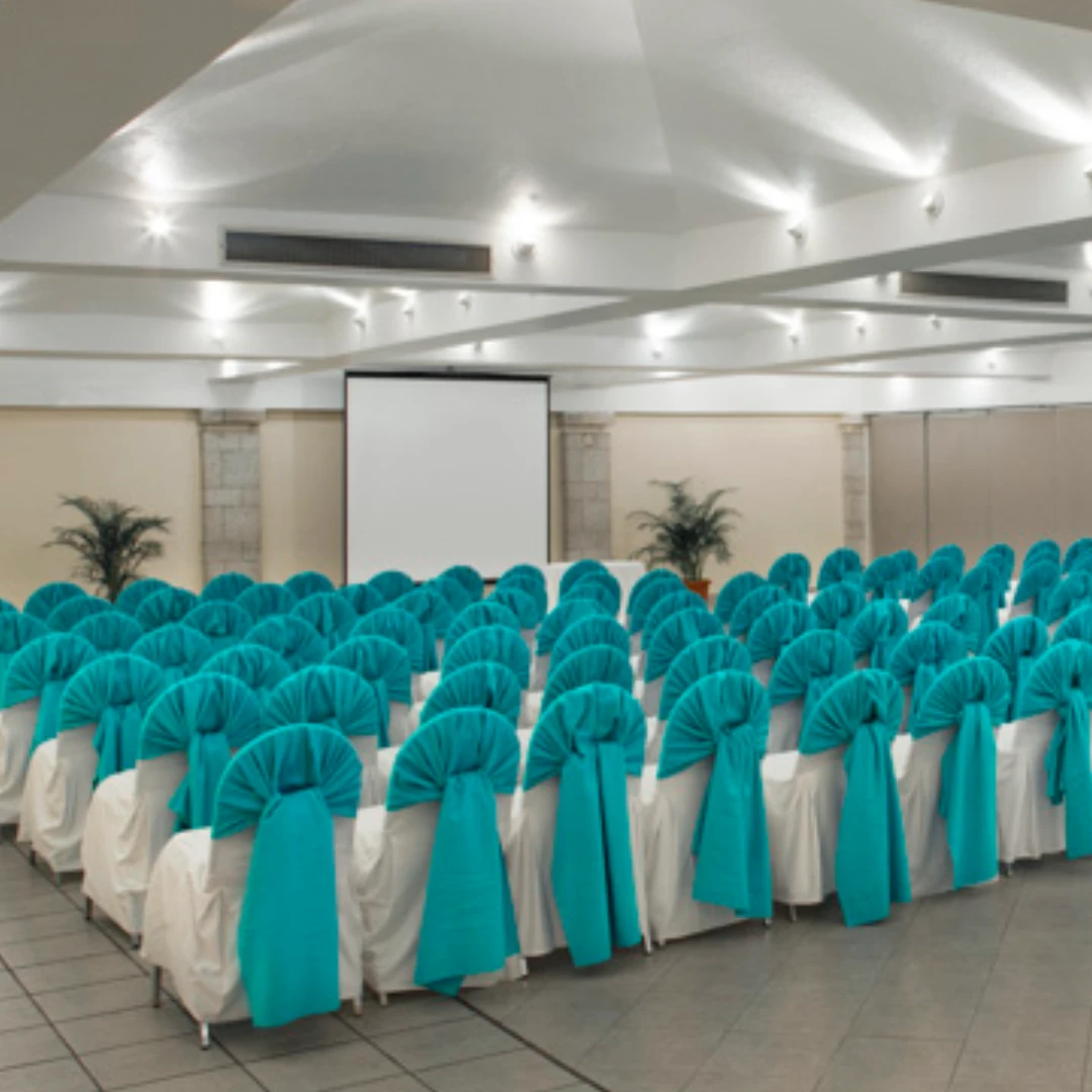 Sala de conferencias con sillas cubiertas de tela turquesa, pantalla de proyecci&oacute;n y plantas decorativas.