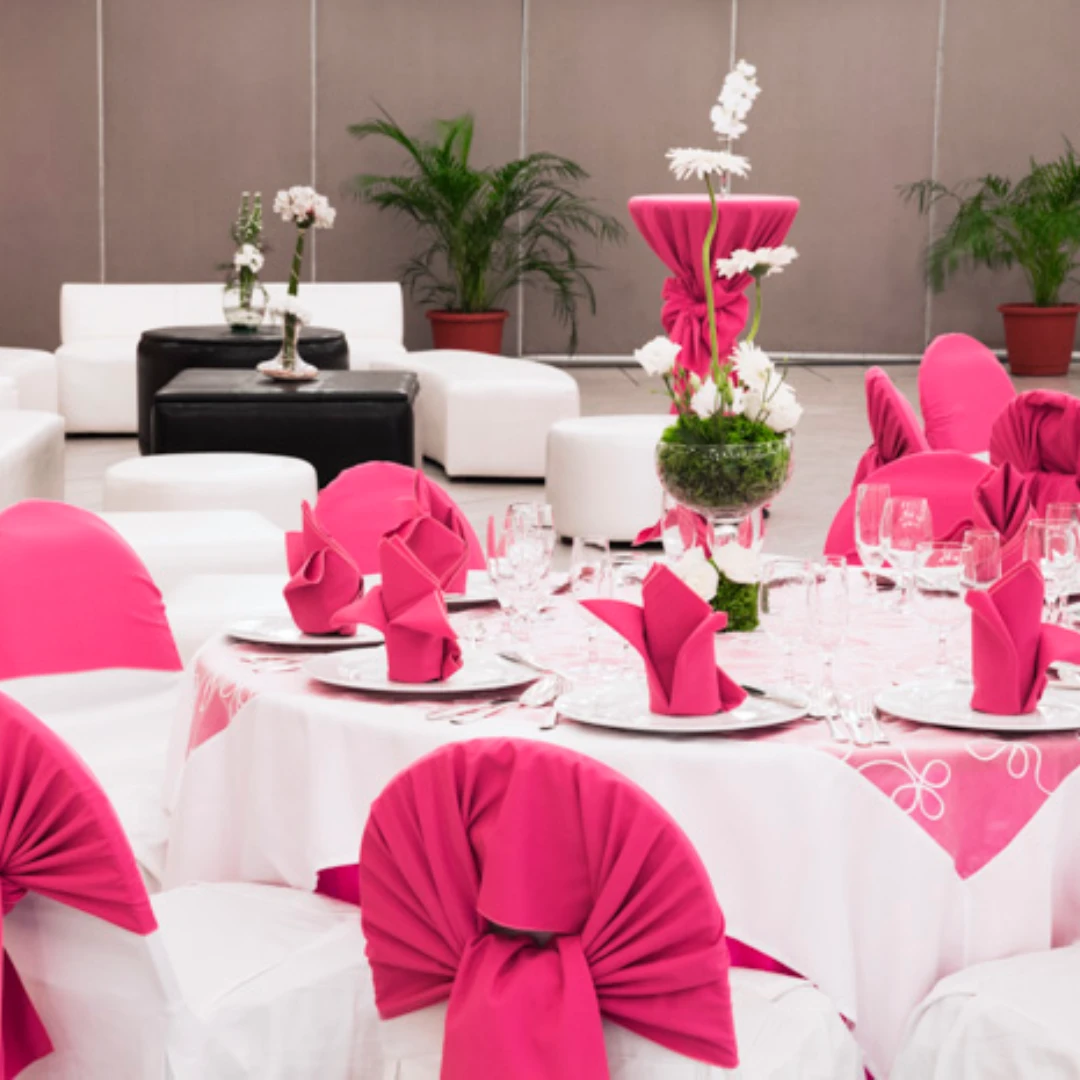 Mesa elegante decorada con manteles y servilletas fucsias, rodeada de sillones blancos y plantas de fondo.