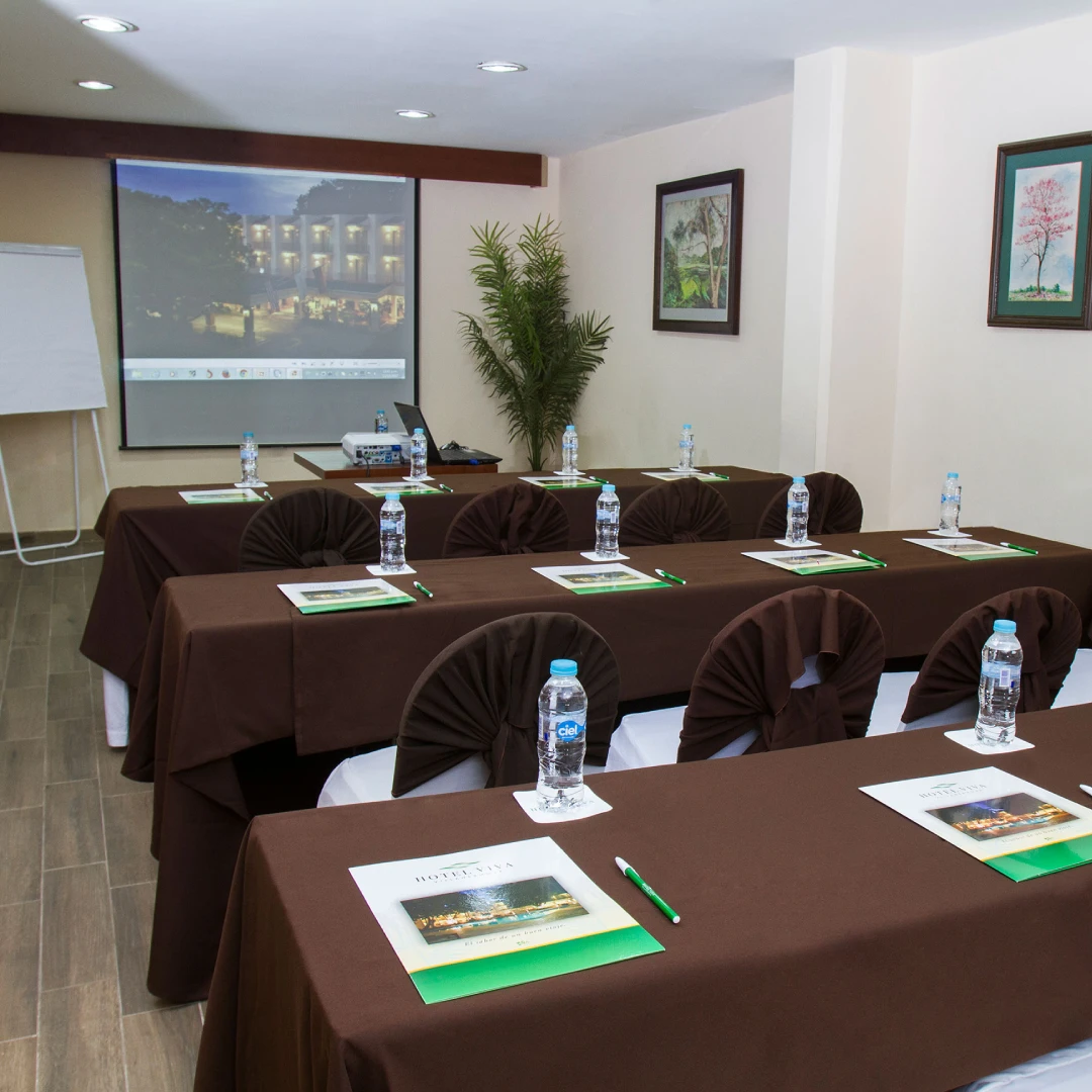 Sala de conferencias con mesas cubiertas de manteles marrones, botellas de agua, bloc de notas y proyector encendido.