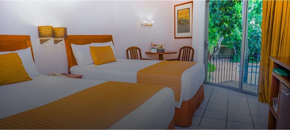 Habitaci&oacute;n de hotel con dos camas, mesa, sillas y una puerta de vidrio que muestra un balc&oacute;n con vegetaci&oacute;n.