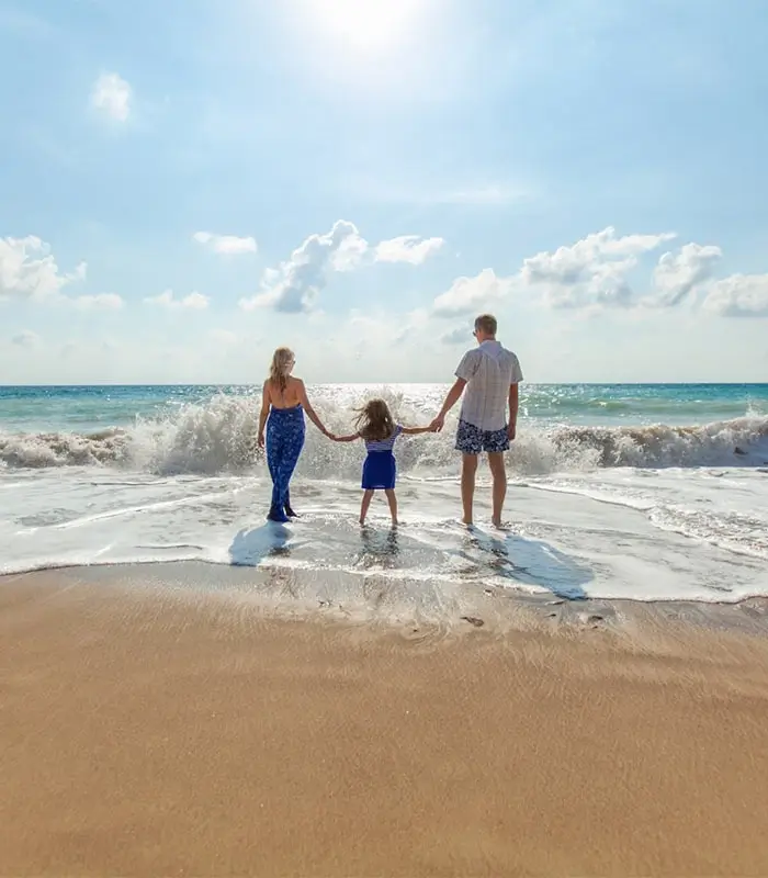 Familia en la playa de pie frente al mar, sosteniendo las manos, con el sol brillando en el cielo azul claro.