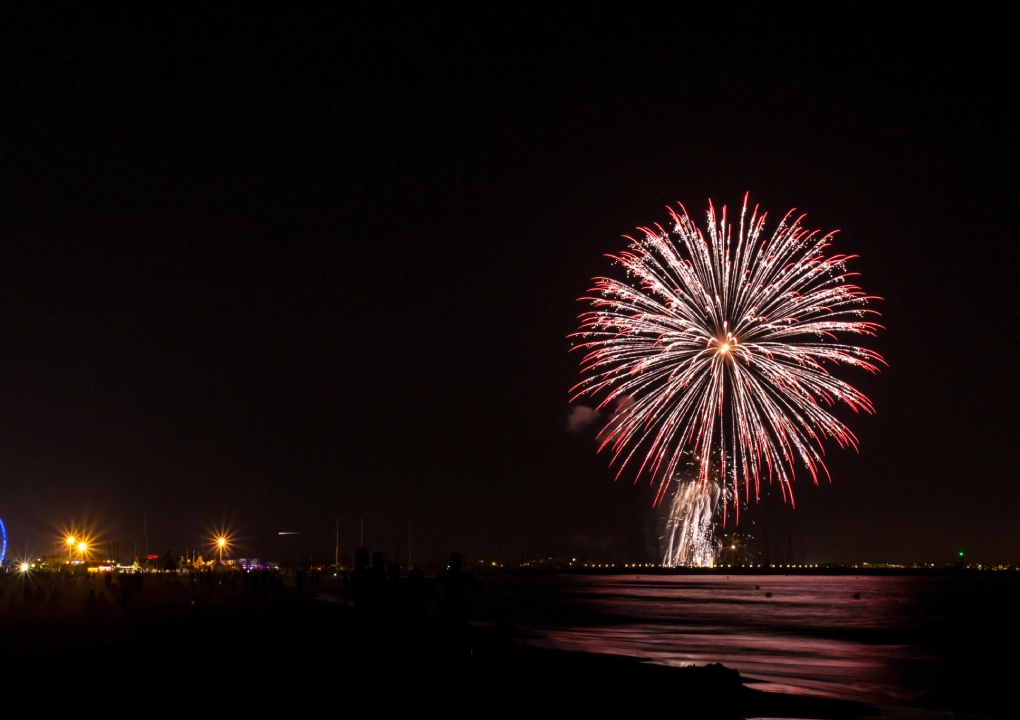 Feu d'artifice color&eacute; illuminant le ciel nocturne au-dessus d'une plage sombre, lumi&egrave;res de f&ecirc;te &agrave; gauche.