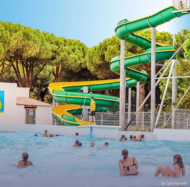 Des gens nagent dans une piscine avec un grand toboggan vert et jaune en arri&egrave;re-plan, entour&eacute; d'arbres.