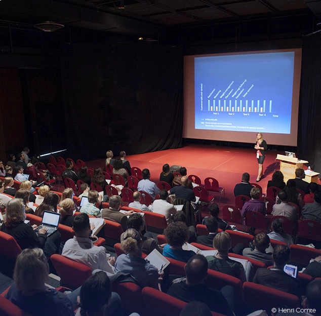 Conf&eacute;rence avec une pr&eacute;sentation graphique, audience assise dans une salle de th&eacute;&acirc;tre.