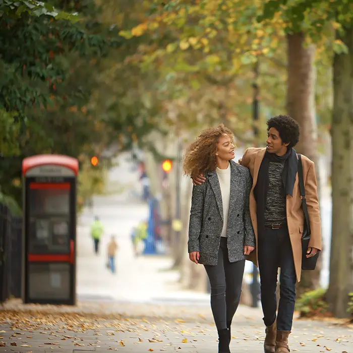 Couple walking on a leafy sidewalk, red phone booth in the background.