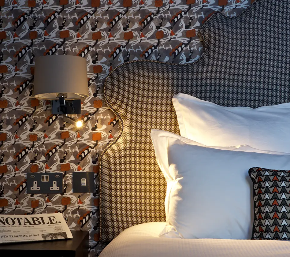 Bedside lamp, patterned wallpaper, pillows, and a newspaper on the nightstand.
