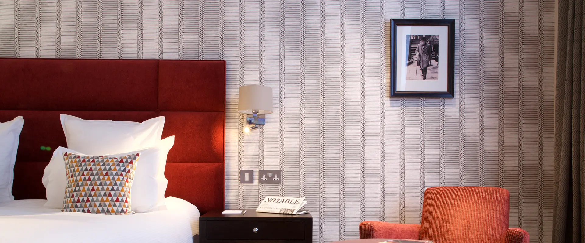 Cozy hotel room with a red bed, patterned wallpaper, and framed photo.
