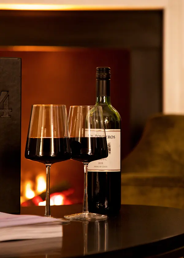 Two wine glasses and a bottle in front of a cozy fireplace.