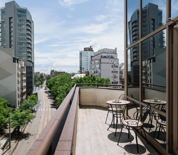 Balcony with tables, chairs, and a city view with tall buildings and trees.