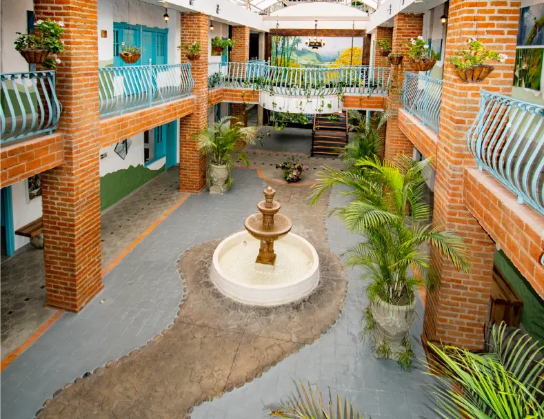 Patio interior con fuente central y balcones de ladrillo, rodeado de plantas y barandales azules.