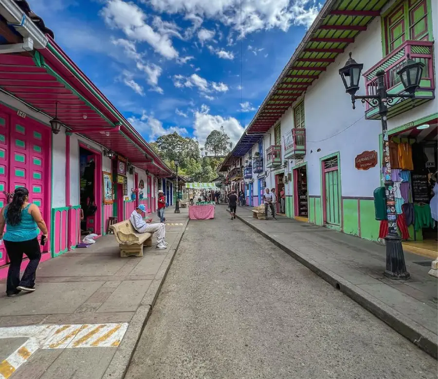 Calle colorida en un pueblo, con personas caminando y un cielo azul con nubes.
