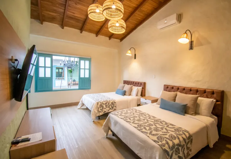 Habitaci&oacute;n luminosa con dos camas, ventana abierta, televisor y l&aacute;mparas colgantes.