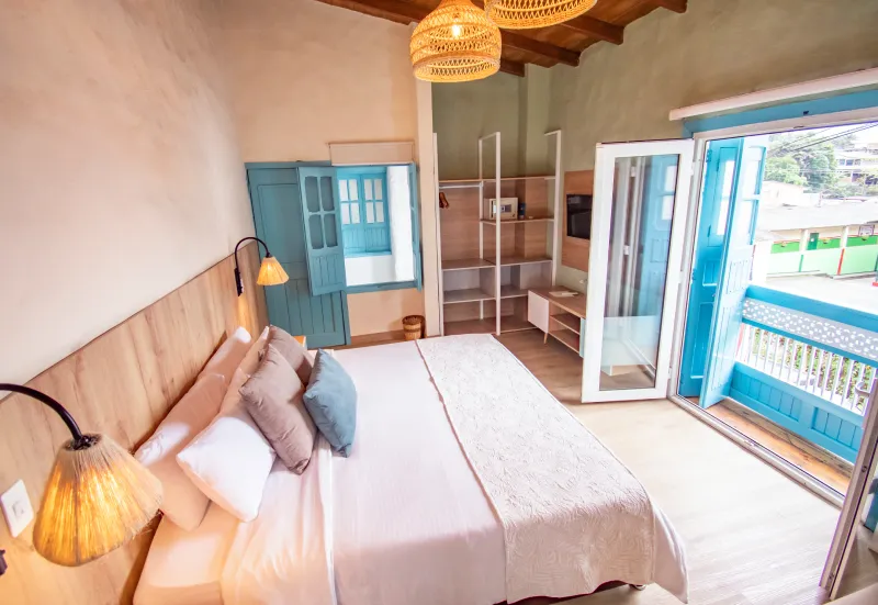 Habitaci&oacute;n con cama, l&aacute;mparas, ventanas azules, puertas abiertas y muebles de madera clara.