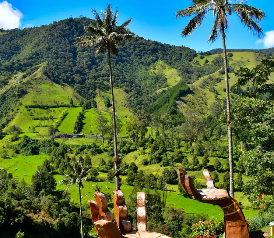 Vista de un valle verde con palmeras y monta&ntilde;as, se ven esculturas de manos hacia el cielo.