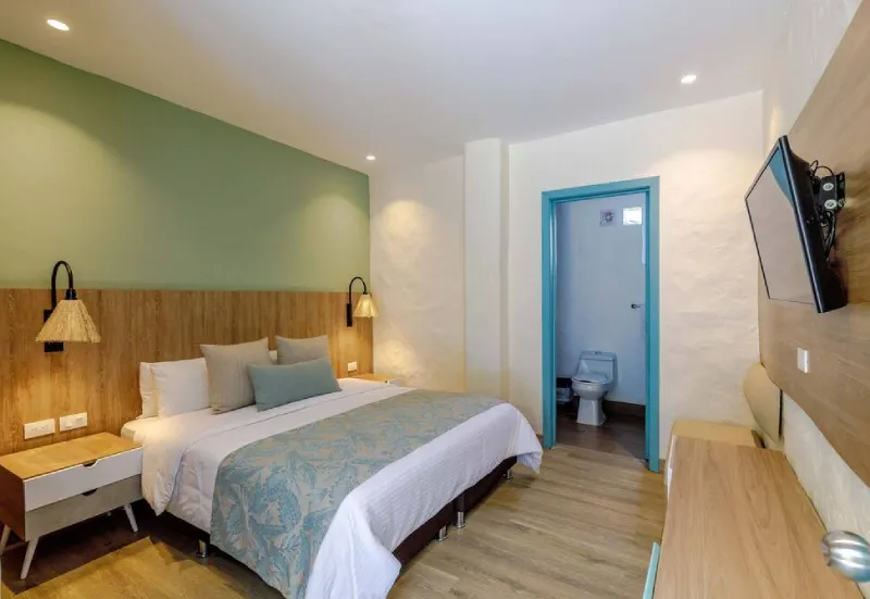 Habitaci&oacute;n con cama doble, l&aacute;mparas, televisor en la pared y puerta a un ba&ntilde;o peque&ntilde;o.