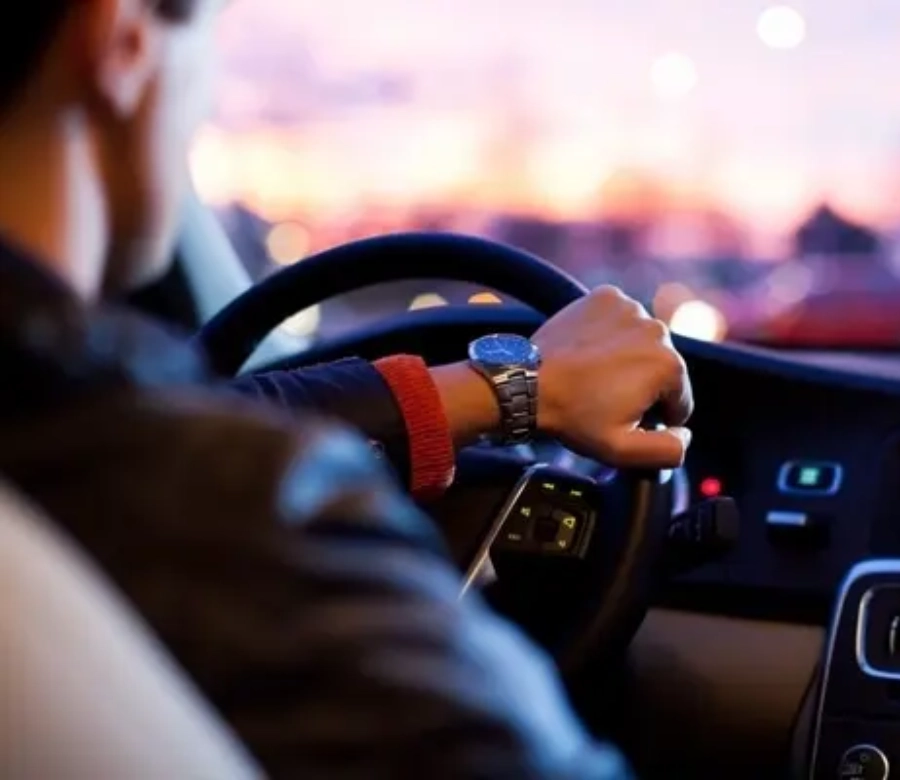 Conductor de perfil al volante de un coche al atardecer, con luces de ciudad al fondo.
