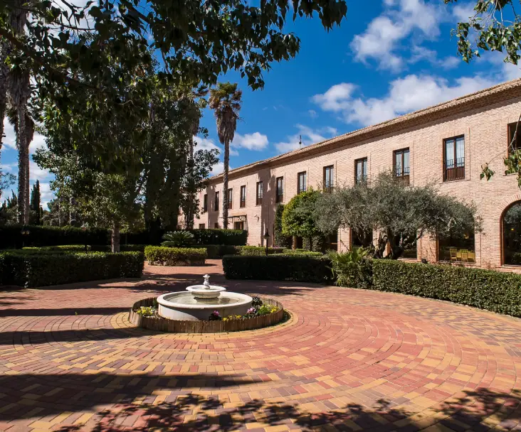 Edificio de ladrillo con jard&iacute;n y fuente central, rodeado de &aacute;rboles bajo un cielo azul.