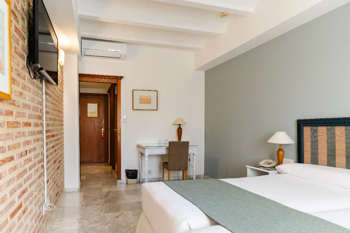 Habitaci&oacute;n de hotel moderna con cama, escritorio y televisi&oacute;n en pared de ladrillo.