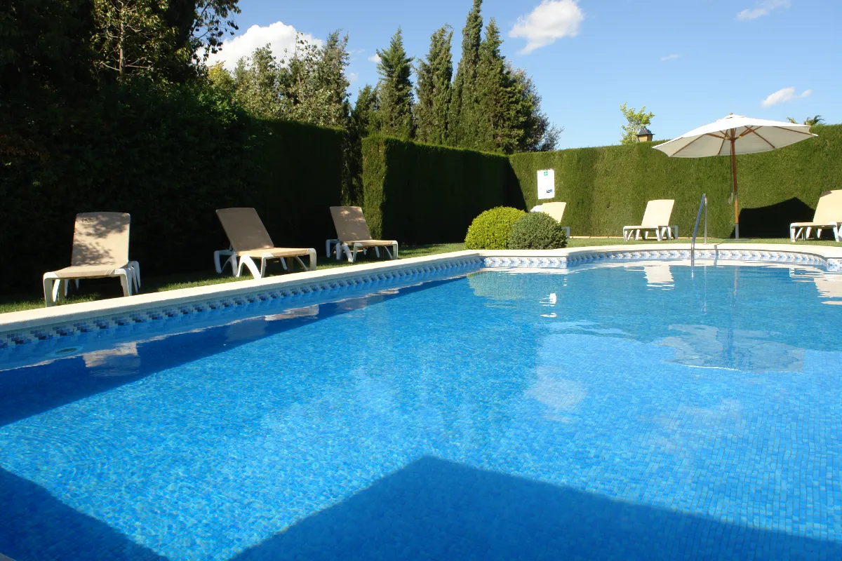 Piscina al aire libre rodeada de tumbonas, setos y &aacute;rboles bajo un cielo azul.