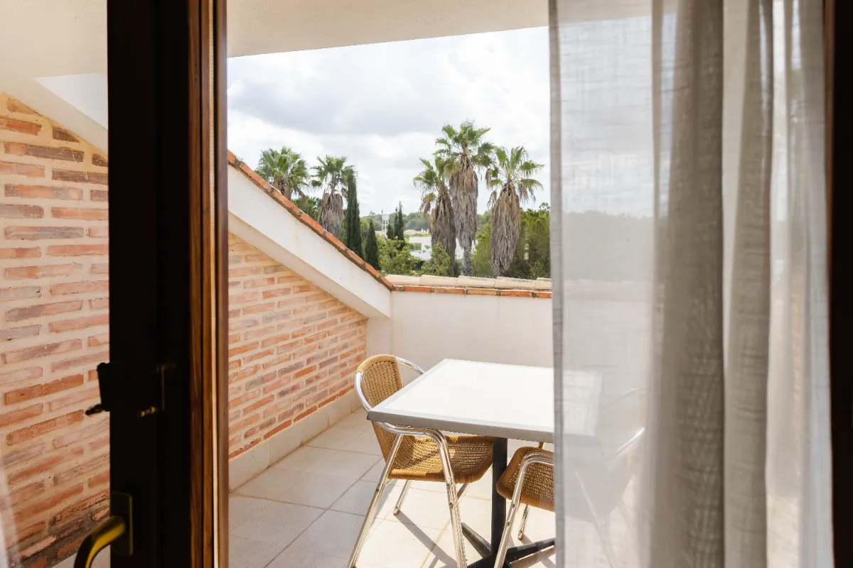 Terraza con mesa, sillas y vista a palmeras, vista desde una puerta de cristal con cortina blanca.