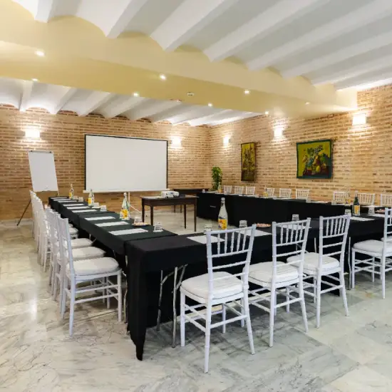 Sala de conferencias con asientos blancos, mesa en U, proyector, pantalla y paredes de ladrillo.