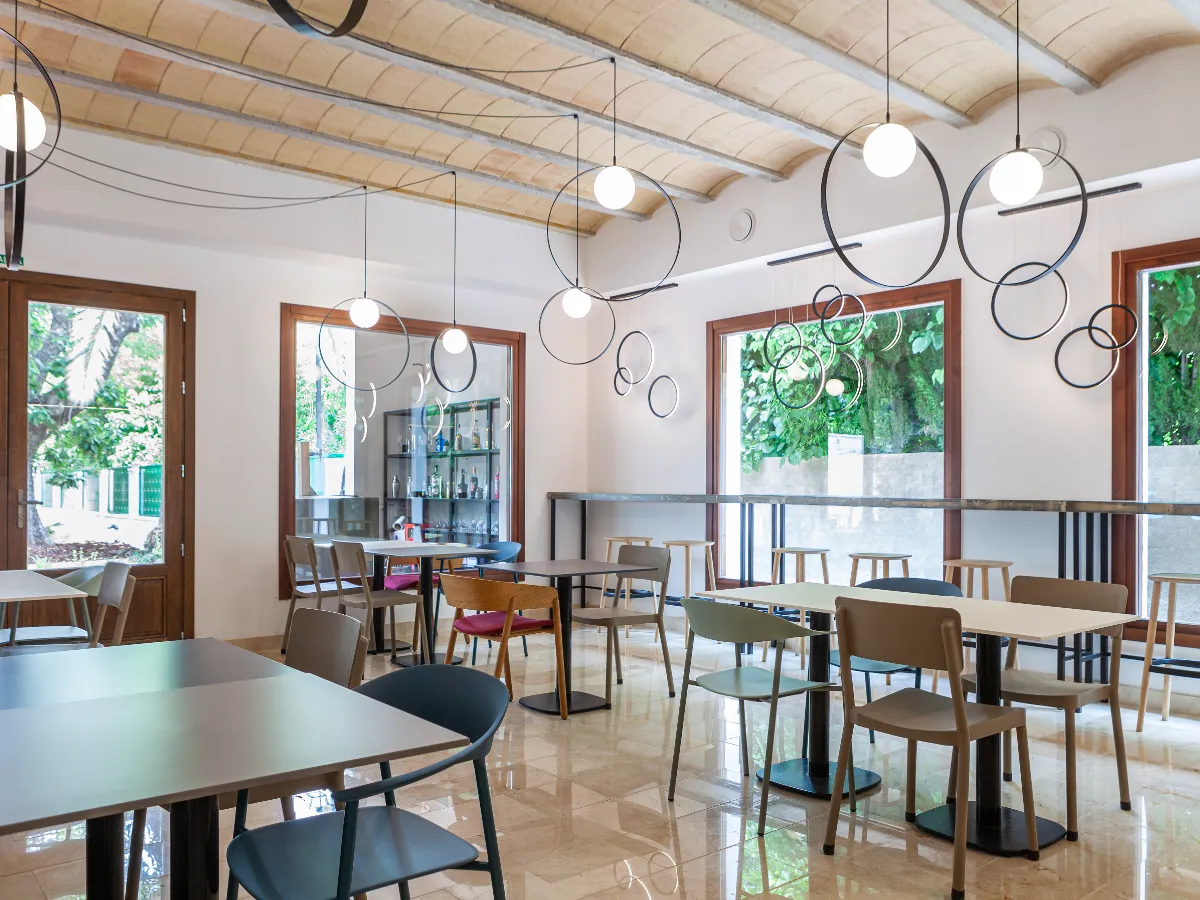 Una cafeter&iacute;a moderna con mesas, sillas y l&aacute;mparas colgantes, vista a trav&eacute;s de grandes ventanas.