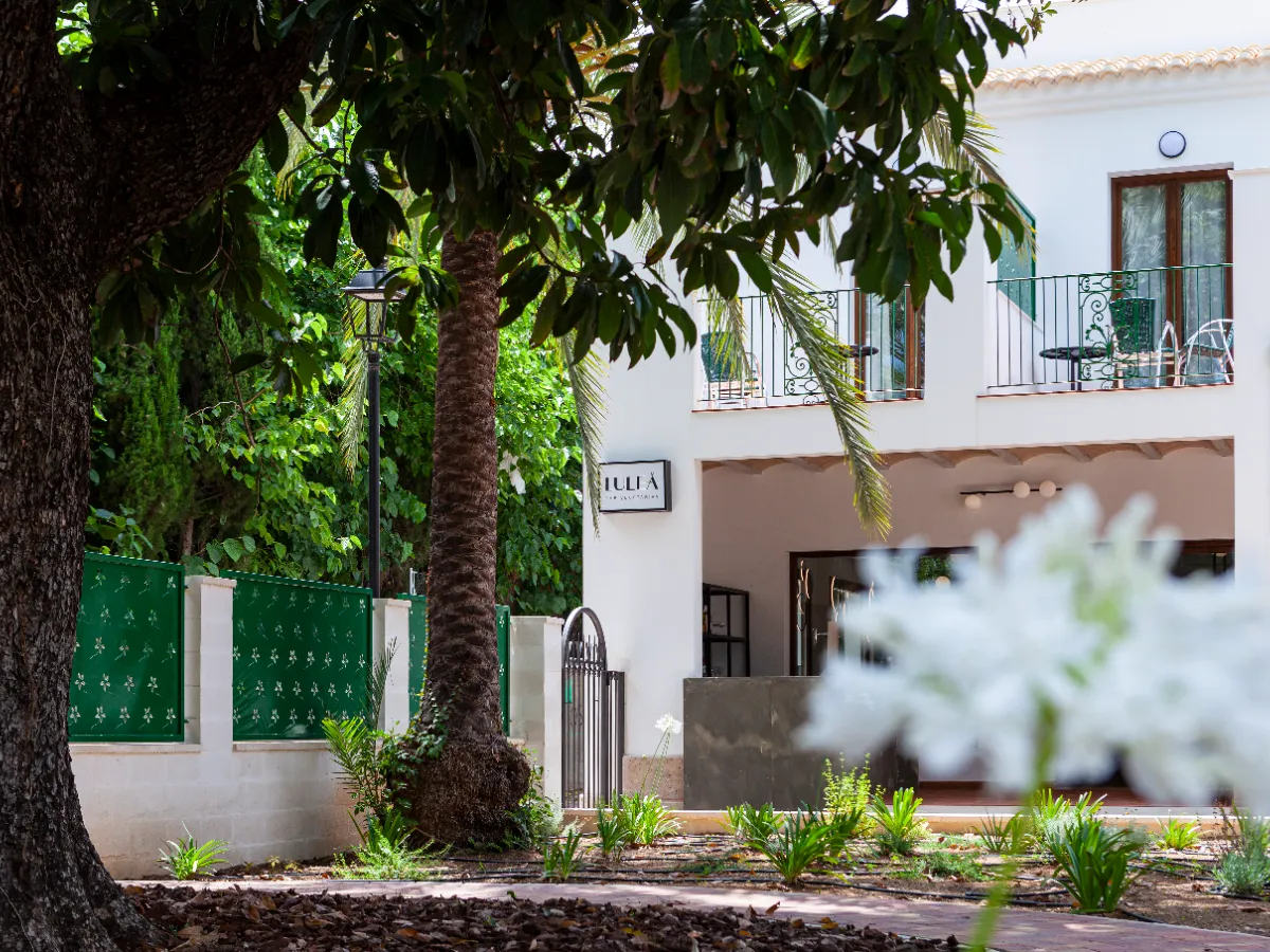 Fachada de hotel rodeada de &aacute;rboles y plantas, con un cartel que dice "Llucia".