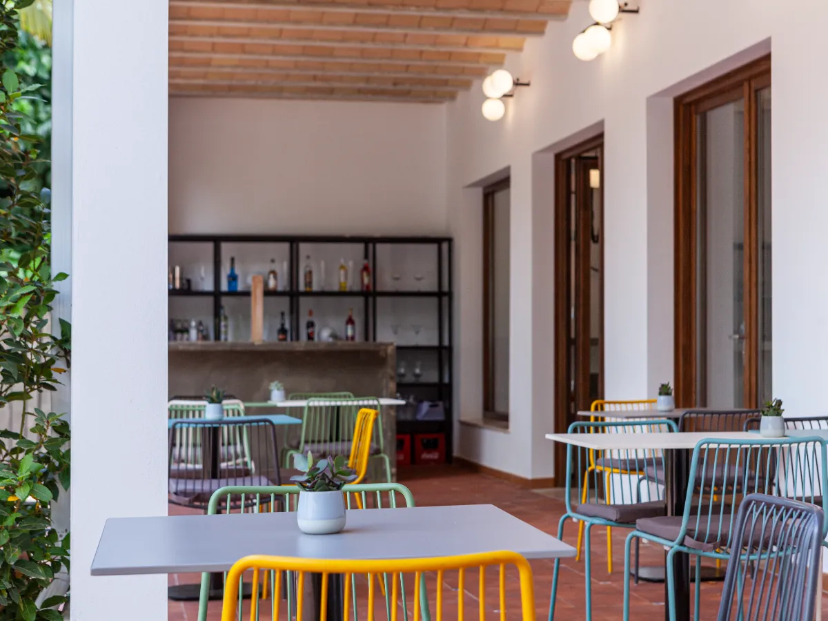 Patio con mesas y sillas de colores, plantas y luces en el techo. Ambiente moderno y acogedor.