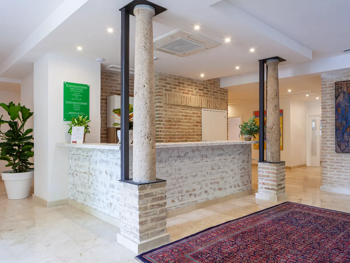 Recepci&oacute;n de hotel con columnas de piedra, plantas y alfombra sobre piso de m&aacute;rmol.