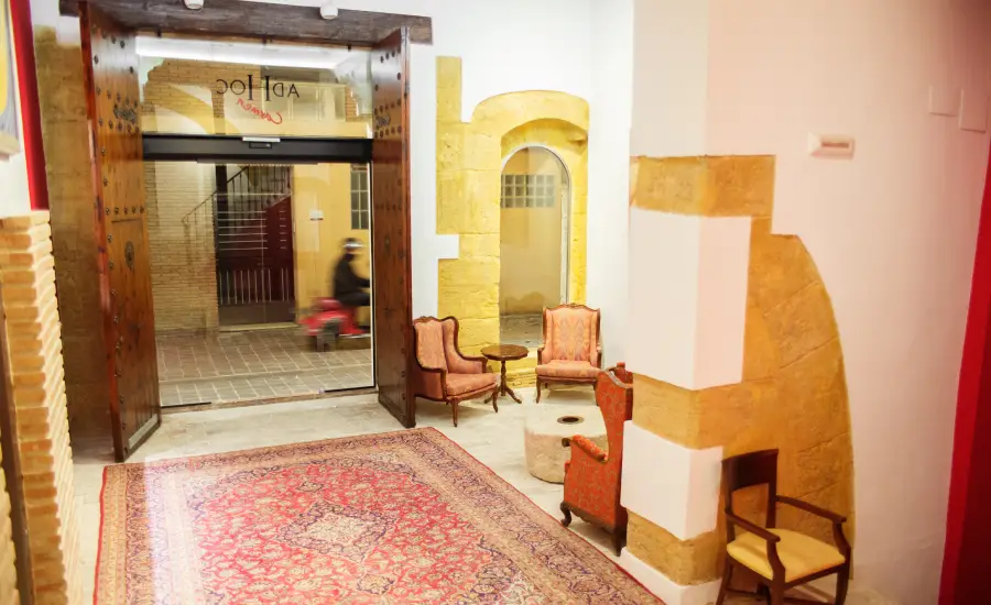 Sala con sillones y alfombra roja, entrada acristalada, vista a la calle y paredes de piedra.