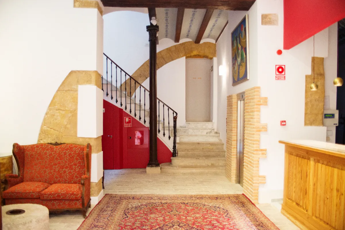 Vest&iacute;bulo con sof&aacute; rojo, alfombra, escalera y mostrador de madera. Decoraci&oacute;n r&uacute;stica.