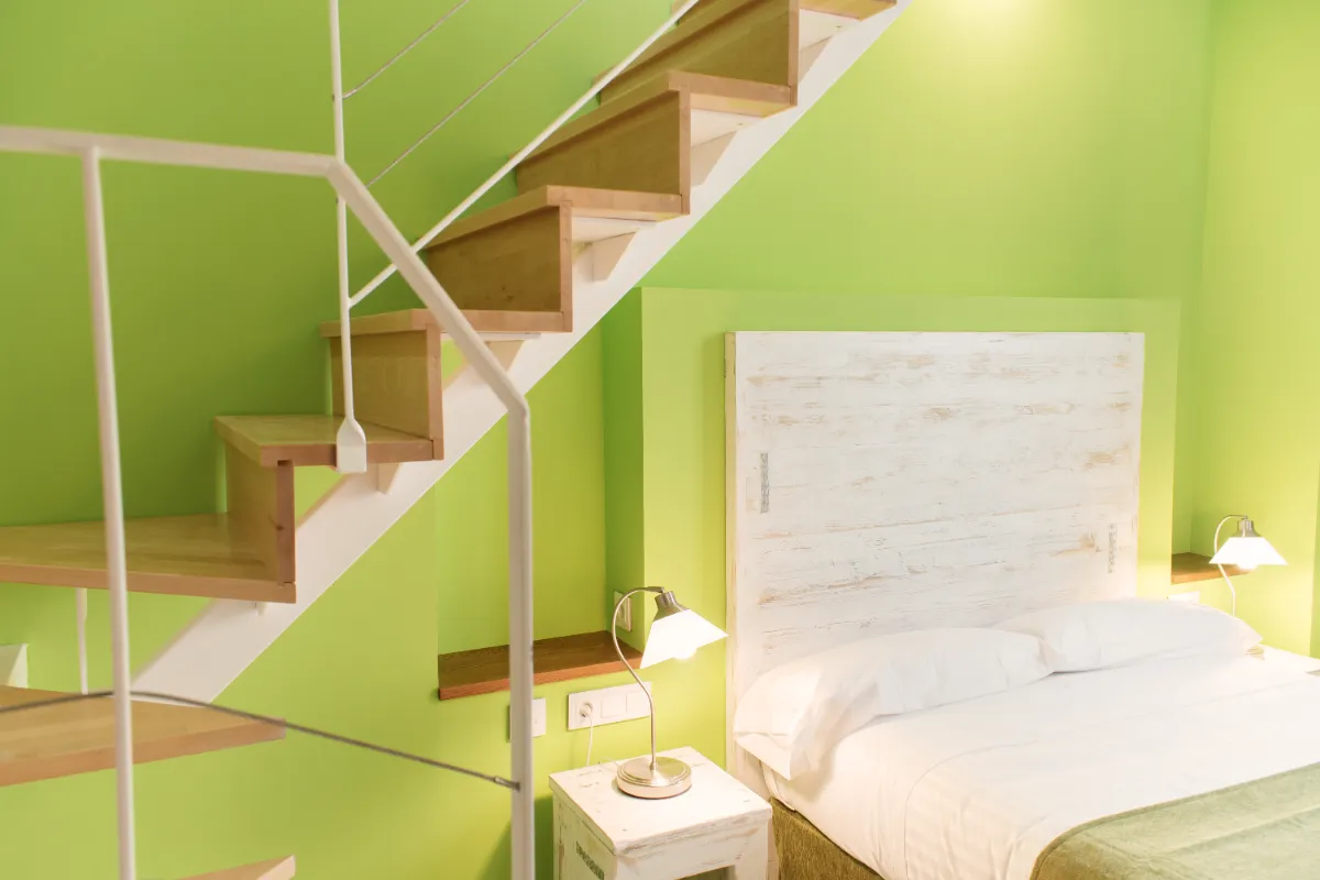 Dormitorio verde con cama blanca y escalera de madera al lado, l&aacute;mpara en mesa de noche.