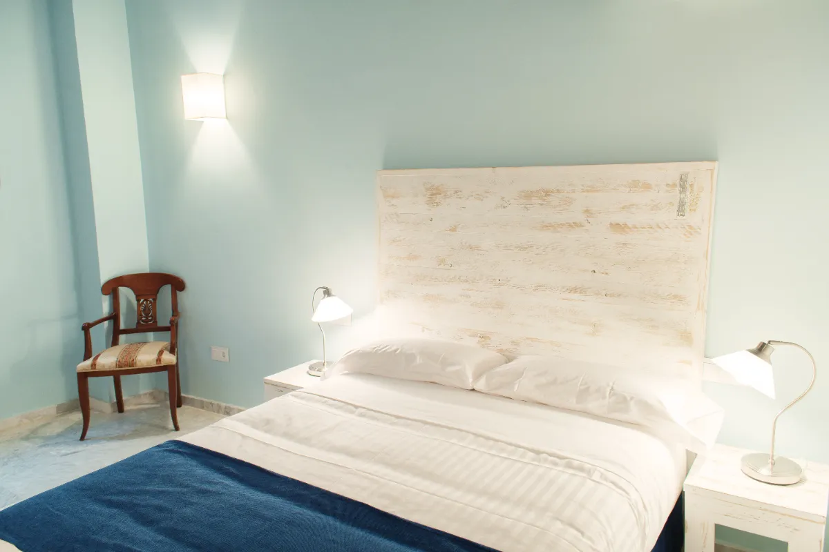 Habitaci&oacute;n luminosa con cama, pared azul, cabecera de madera blanca y l&aacute;mparas de noche.