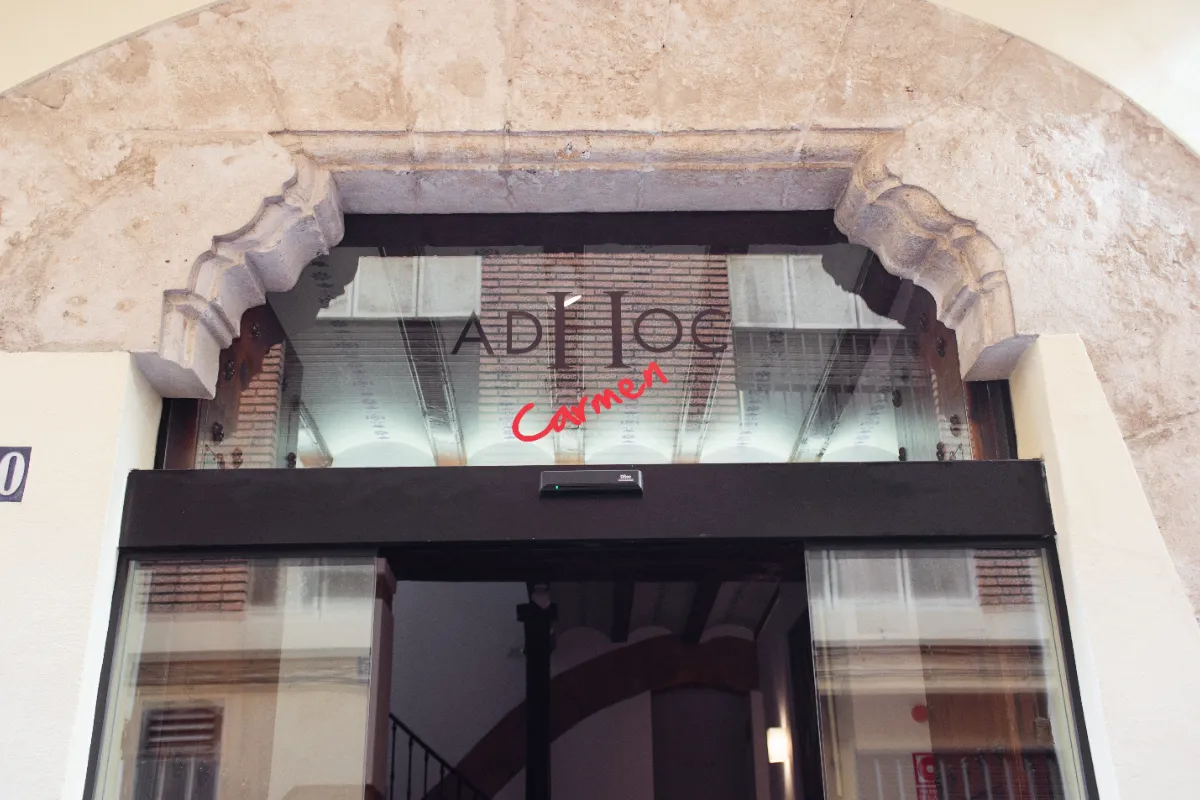 Puerta de cristal con el letrero "AD Hoc Carmen" sobre un arco de piedra.
