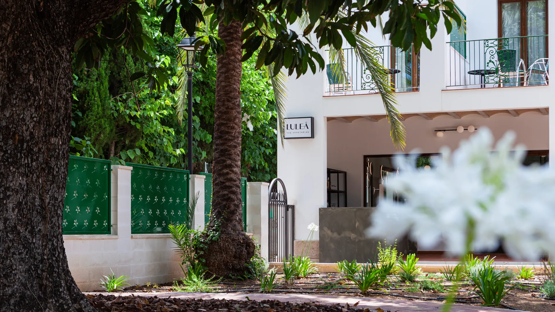 Exteriores de un restaurante rodeado de &aacute;rboles y flores, con un cartel visible en la fachada.