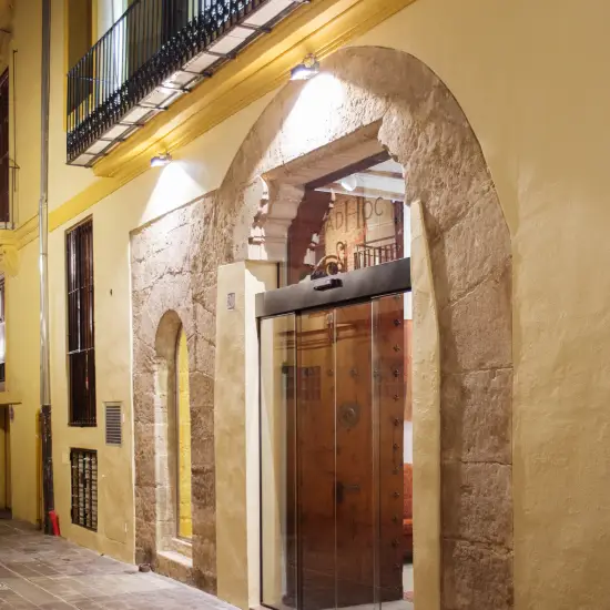 Puerta de vidrio en edificio antiguo con arco de piedra y luces exteriores, durante la noche.