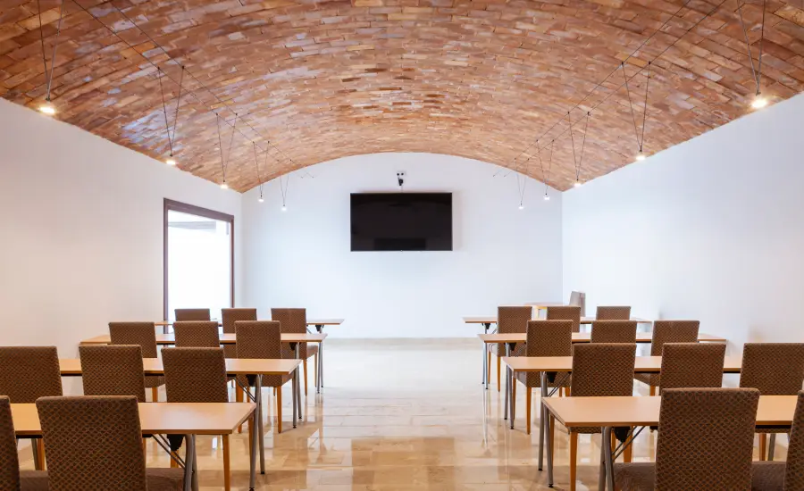 Sala de conferencias con techo de ladrillo, mesas, sillas y pantalla grande al fondo.